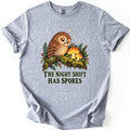 The Night Shift Has Spores Comfort Colors T-shirt in Sport Grey, laid flat. A sleepy brown owl perches on a mossy branch beside two glowing orange mushrooms, with ferns, red berries and small stars, above the words The Night Shift Has Spores in dark green. Shown on white.