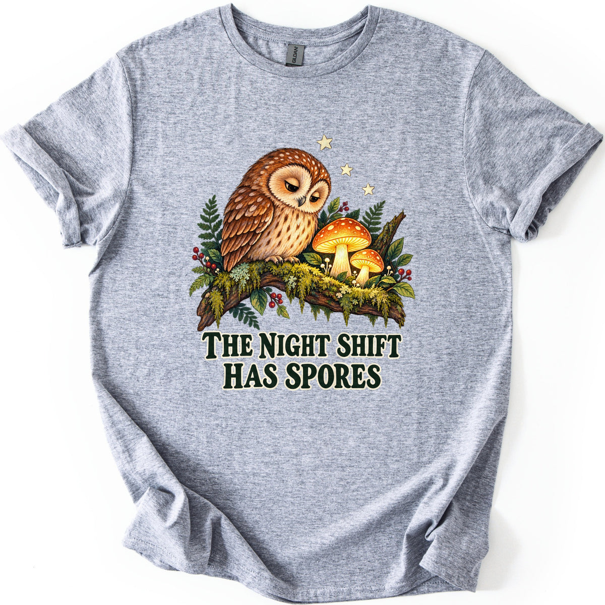 The Night Shift Has Spores Comfort Colors T-shirt in Sport Grey, laid flat. A sleepy brown owl perches on a mossy branch beside two glowing orange mushrooms, with ferns, red berries and small stars, above the words The Night Shift Has Spores in dark green. Shown on white.