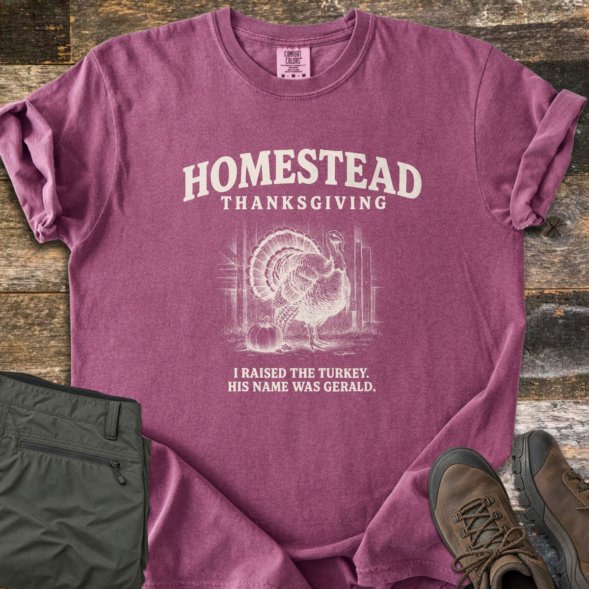 Homestead Thanksgiving Comfort Colors T-shirt in Berry, laid flat. Cream print of a fanned-tail turkey by a barn door and pumpkin, under the words Homestead Thanksgiving, above I Raised the Turkey. His Name Was Gerald. On weathered wood with olive pants and brown hiking boots.