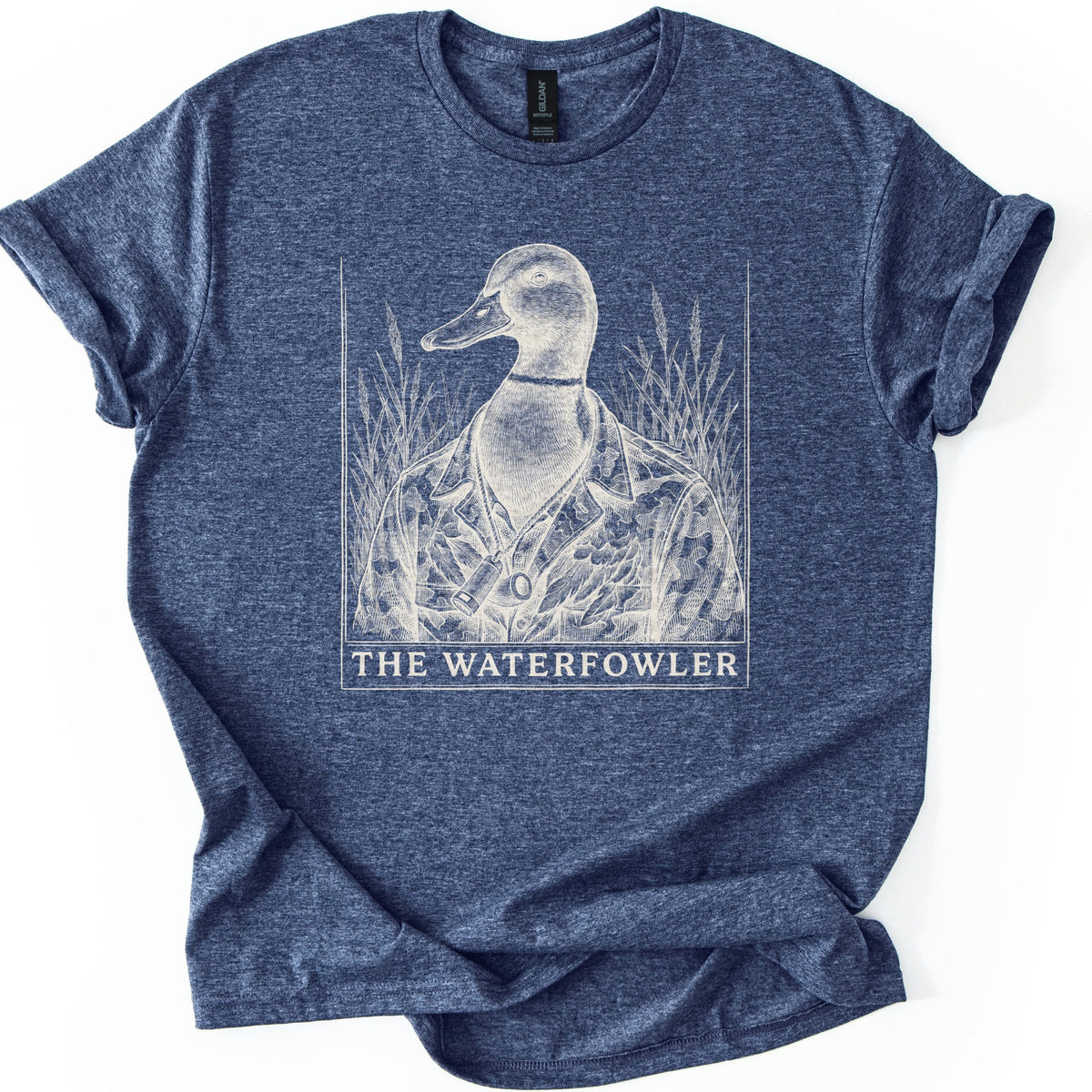 The Waterfowler Comfort Colors T-shirt in Heather Navy, laid flat. A white engraving-style drawing of a mallard duck in a camo shirt with a duck call on a lanyard, set in tall marsh grass inside a thin frame, above the words THE WATERFOWLER. Sleeves cuffed, on white.