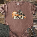 Dune Drifter Comfort Colors T-shirt in Espresso, laid flat. A rider in a helmet drifts an orange and black ATV through a spray of orange sand above the words Dune Drifter. Sleeves are rolled on a wood plank surface beside folded olive cargo pants and brown hiking boots.
