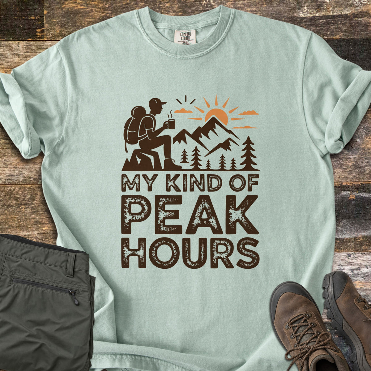 My Kind Of Peak Hours Comfort Colors T-shirt in Bay, laid flat. A dark brown hiker with a backpack sits on a rock with a steaming mug, facing mountains, pines and an orange sun, above the words My Kind Of Peak Hours. Olive hiking pants and brown boots lie on weathered wood.