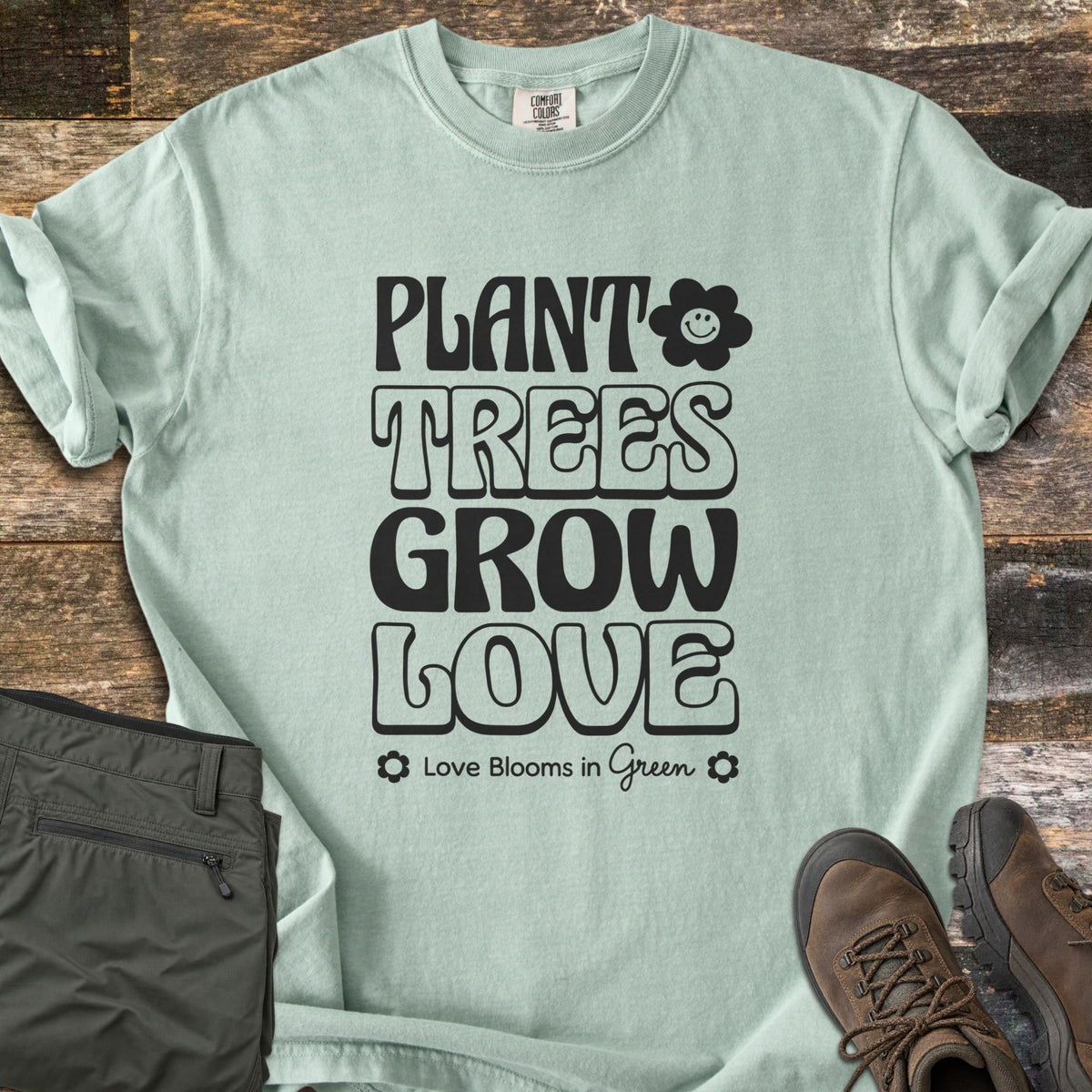 Plant Trees Grow Love Comfort Colors T-shirt in Bay, laid flat. Black retro bubble letters read Plant Trees Grow Love beside a smiling daisy, above Love Blooms in Green. Sleeves cuffed on weathered wood with folded olive hiking pants and brown hiking boots.