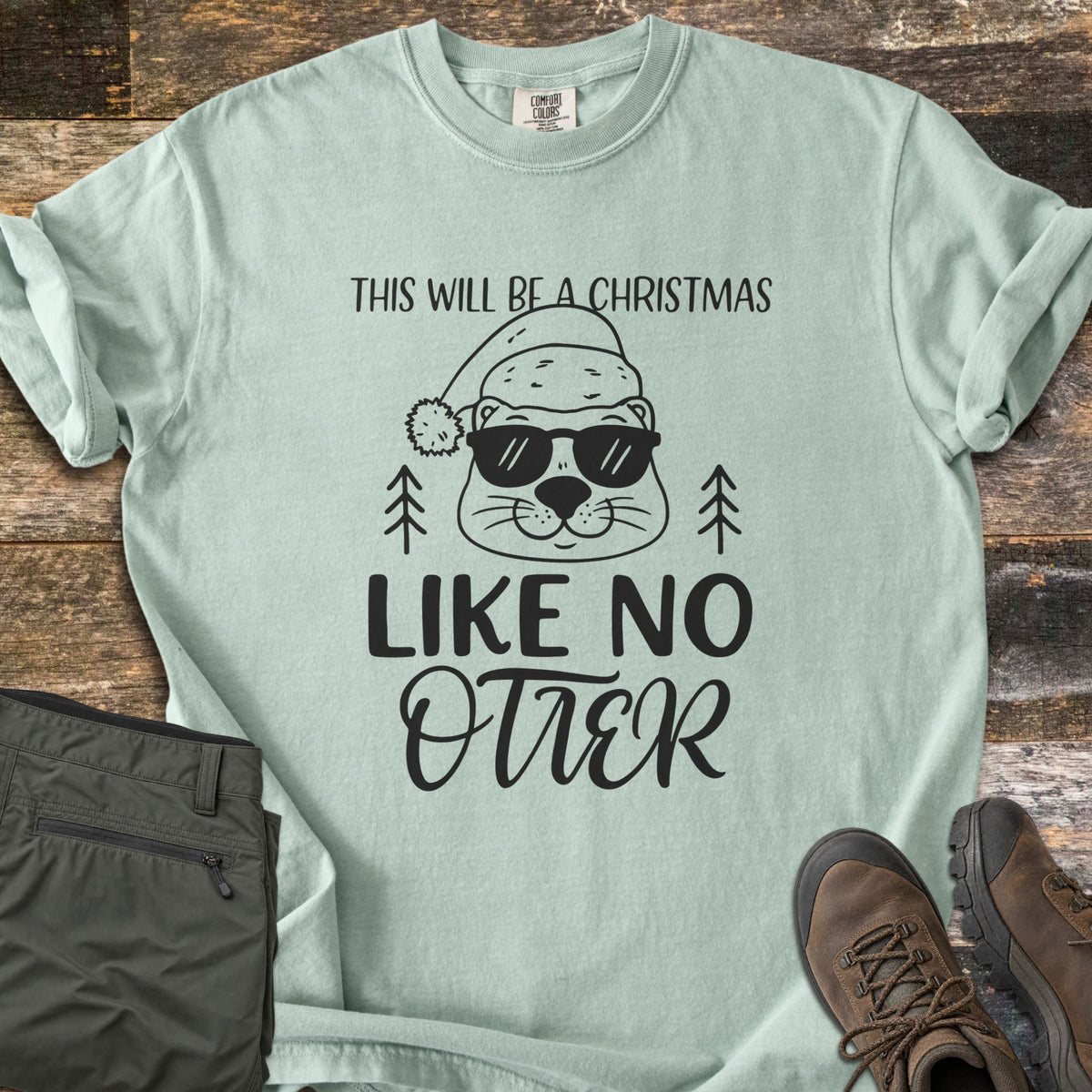 Christmas Like No Otter Comfort Colors T-shirt in Bay, laid flat. A cartoon otter in a Santa hat and sunglasses sits between two pine trees, with This Will Be a Christmas above and Like No Otter below, in black. Olive cargo pants and brown hiking boots lie on weathered wood.