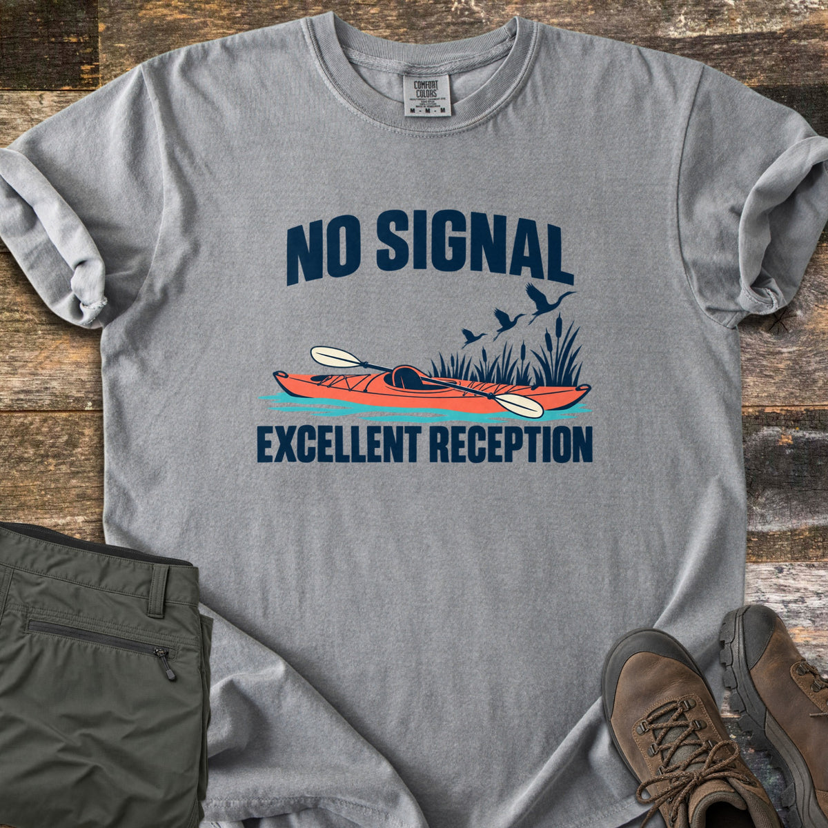 Excellent Reception Comfort Colors T-shirt in Grey, laid flat. The words No Signal arch over an orange kayak with a paddle among reeds and flying birds, above the words Excellent Reception. Olive pants and brown hiking boots sit on a weathered wood surface.