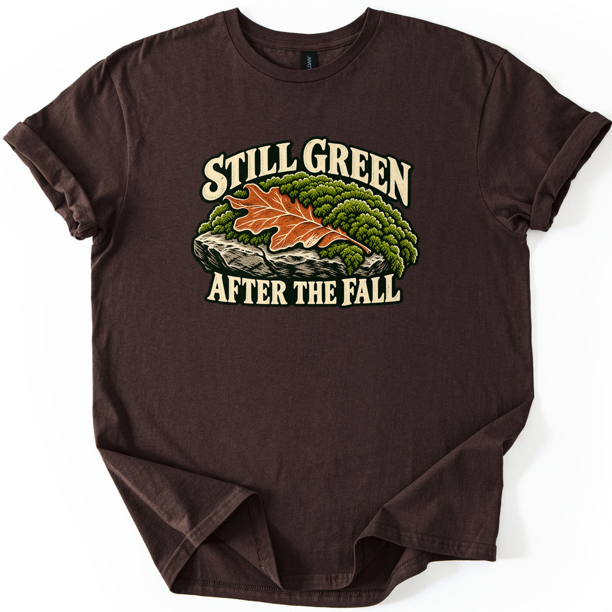 Still Green After the Fall Comfort Colors T-shirt in Dark Chocolate, laid flat. An orange oak leaf rests on green moss atop a gray rock ledge, between cream words reading Still Green above and After the Fall below. The sleeves are cuffed, on a white background.