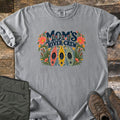 Moms River Crew Comfort Colors T-shirt in Grey, laid flat. The words Mom's River Crew in dark navy letters above red, yellow and green kayaks, framed by coral flowers, cattails and leaves. Olive hiking pants and brown boots lie beside it on weathered wood.