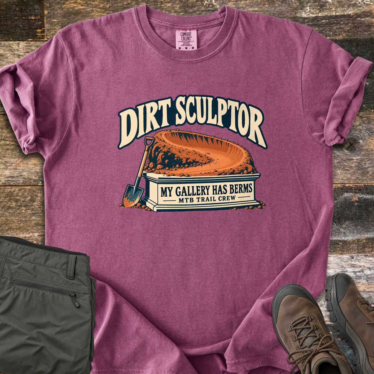 Dirt Sculptor Comfort Colors T-shirt in Berry, laid flat. The words Dirt Sculptor arch over a dirt berm on a stone pedestal reading My Gallery Has Berms, MTB Trail Crew, with a shovel leaning on it. Olive pants and brown hiking boots lie on wood planks.
