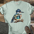 Kingfisher Waterfall Comfort Colors T-shirt in Bay, laid flat. A painted belted kingfisher perches on a branch, a waterfall pouring into a turquoise pool among rocks and pine trees within its body. The shirt lies on weathered wood beside olive pants and brown hiking boots.