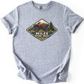 Miles Before Emails Comfort Colors T-shirt in Sport Grey, laid flat. A diamond-shaped patch design shows an orange ATV on rocks with pine trees and mountains, above the words Miles Before Emails. The shirt lies on a white background with its sleeves cuffed.
