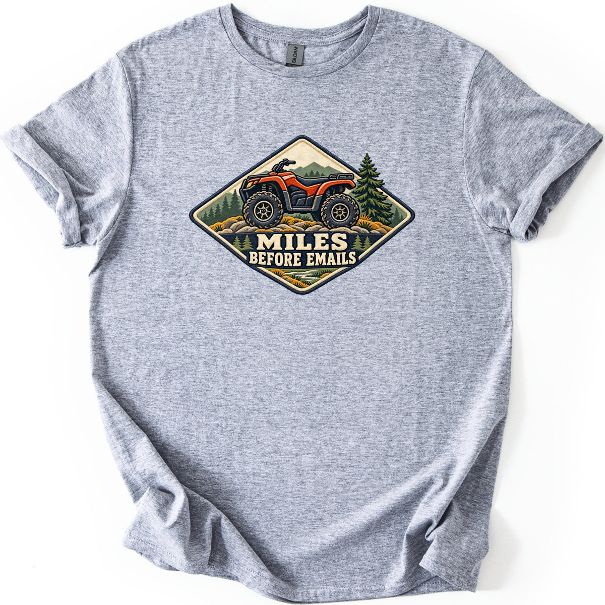 Miles Before Emails Comfort Colors T-shirt in Sport Grey, laid flat. A diamond-shaped patch design shows an orange ATV on rocks with pine trees and mountains, above the words Miles Before Emails. The shirt lies on a white background with its sleeves cuffed.