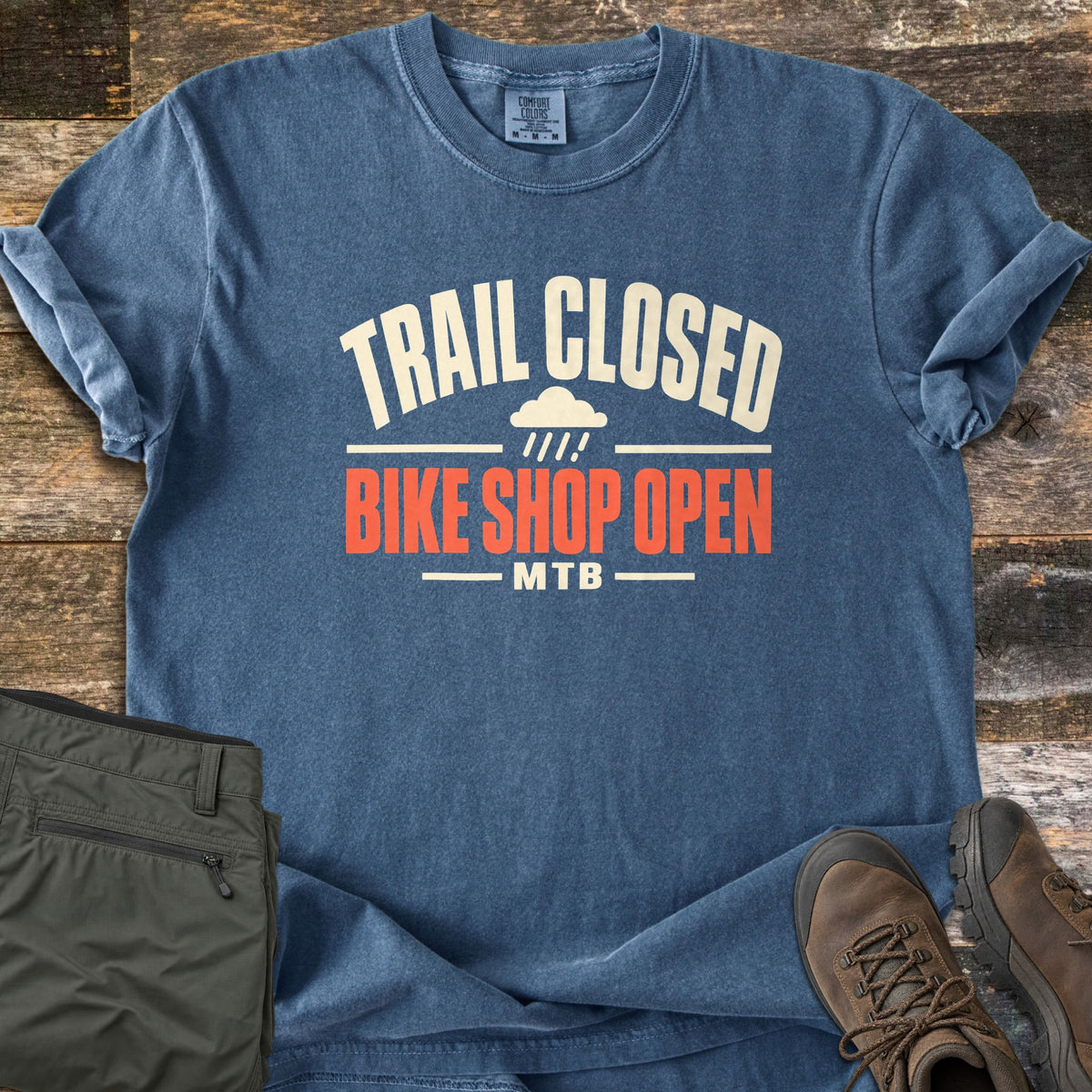 Trail Closed Bike Shop Open Comfort Colors T-shirt in Denim, laid flat. Cream arched text reads Trail Closed above a small rain cloud, with Bike Shop Open in orange-red and MTB below. Sleeves are rolled on weathered wood planks beside folded olive hiking pants and brown boots.