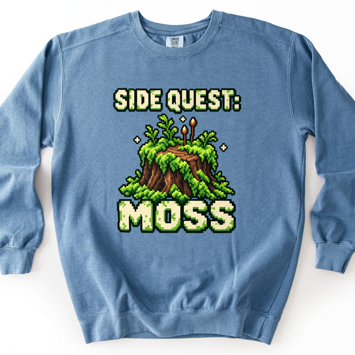 Side Quest Moss Comfort Colors crewneck sweatshirt in Blue Jean, laid flat. Pixel-art tree stump covered in green moss and small ferns, with two orange-tipped sprouts and sparkles, between the words Side Quest: and Moss in mossy cream pixel letters. Shown on a white background.