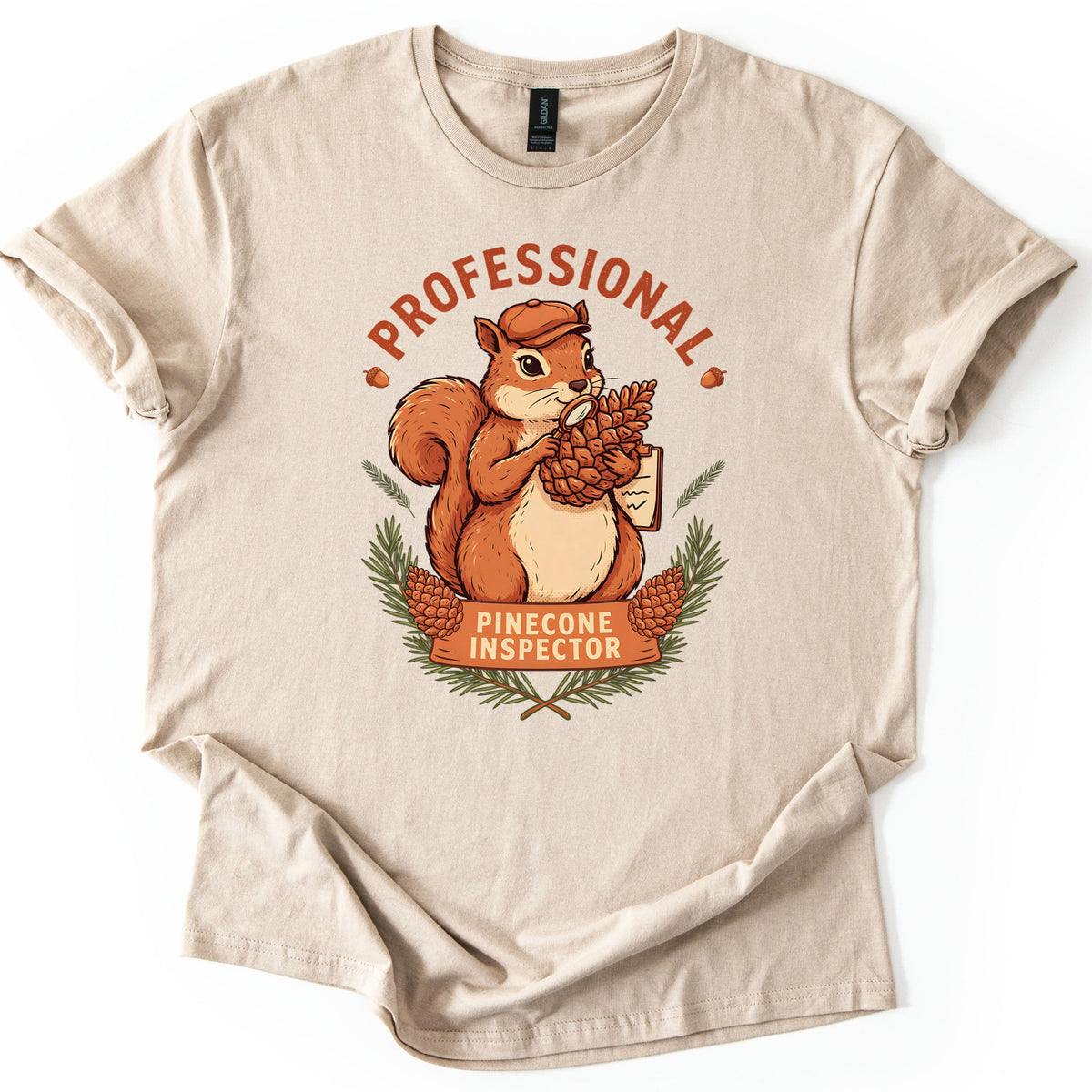 Professional Pinecone Inspector Comfort Colors T-shirt in Sand, laid flat. A squirrel in a cap inspects a pinecone with a magnifying glass under the arched word Professional in rust red, above a banner reading Pinecone Inspector framed by pine branches. Sleeves are cuffed on a white surface.