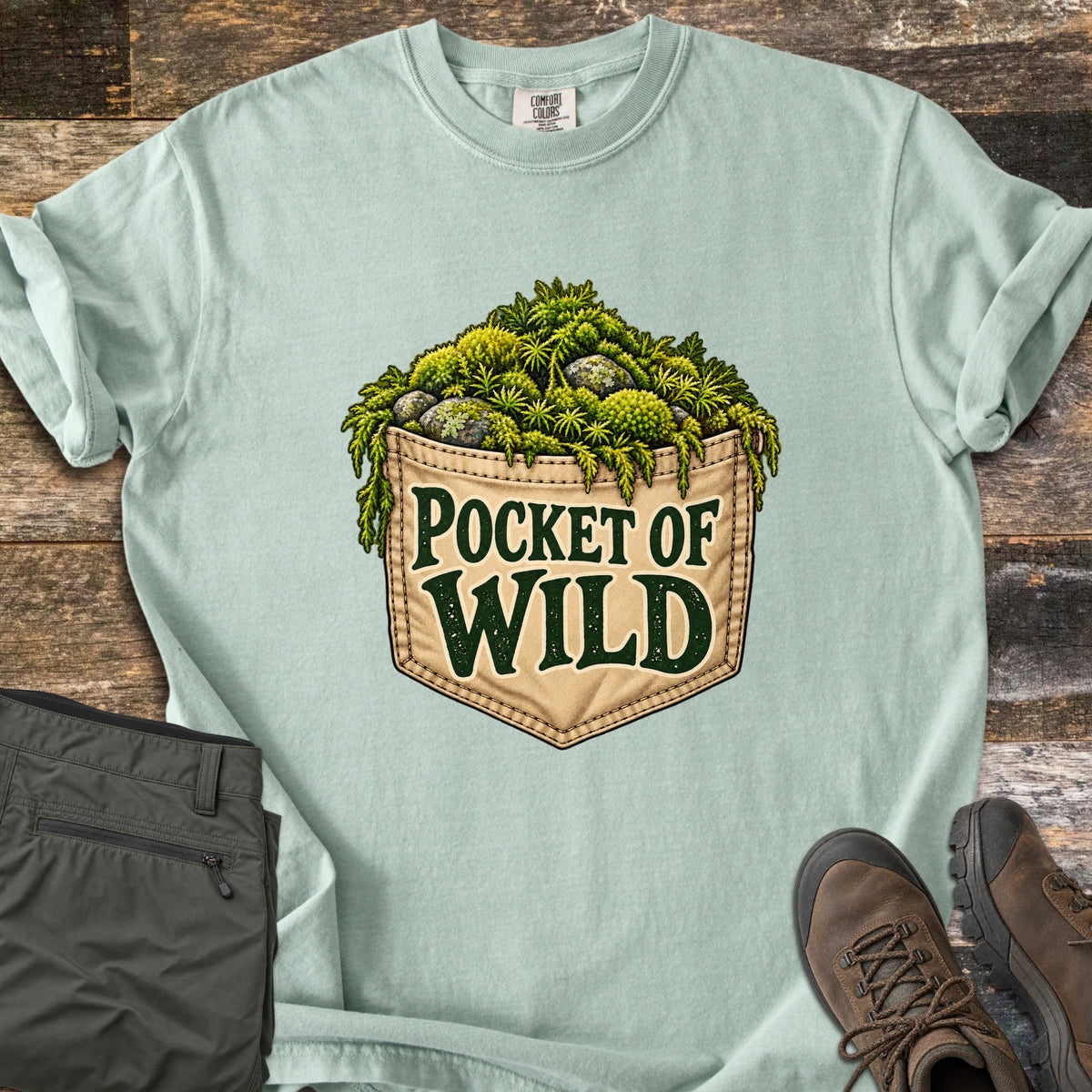 Pocket of Wild Comfort Colors T-shirt in Bay, laid flat. A tan stitched jeans pocket overflowing with green moss and lichen-covered stones, printed with the words Pocket of Wild. Shown on weathered wood with folded olive hiking pants and brown hiking boots.