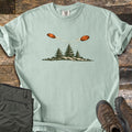 Crossing Clays Comfort Colors T-shirt in Bay, laid flat. Two orange clay targets fly across each other, trailing streaks, above three pine trees on a grassy hill, with no words. The shirt lies on weathered wood beside folded olive pants and brown hiking boots.