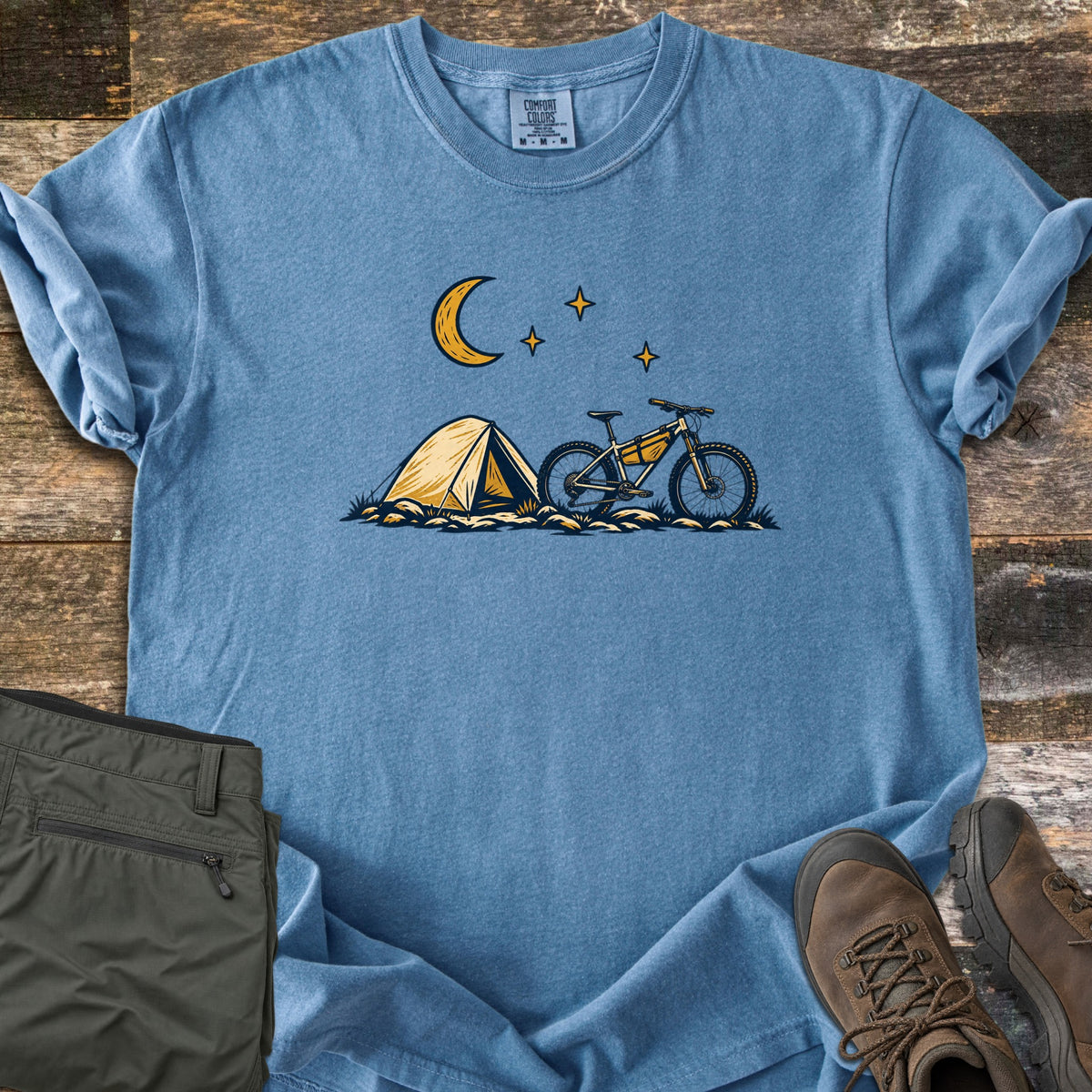 Bikepacking Under the Stars Comfort Colors T-shirt in Blue Jean, laid flat. A yellow tent and a mountain bike with a frame bag sit on grassy ground under a crescent moon and three stars, with no words. Olive hiking pants and brown boots lie beside it on weathered wood planks.