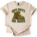 Off Duty on Moss Comfort Colors T-shirt in Sand, laid flat. A worn brown leather hiking boot overgrown with green moss and ferns sits on mossy stones between the words OFF DUTY and ON MOSS in dark green letters outlined in cream. The shirt lies on a white surface.