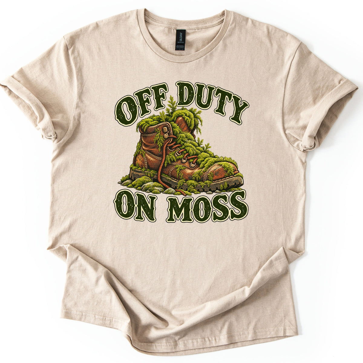 Off Duty on Moss Comfort Colors T-shirt in Sand, laid flat. A worn brown leather hiking boot overgrown with green moss and ferns sits on mossy stones between the words OFF DUTY and ON MOSS in dark green letters outlined in cream. The shirt lies on a white surface.
