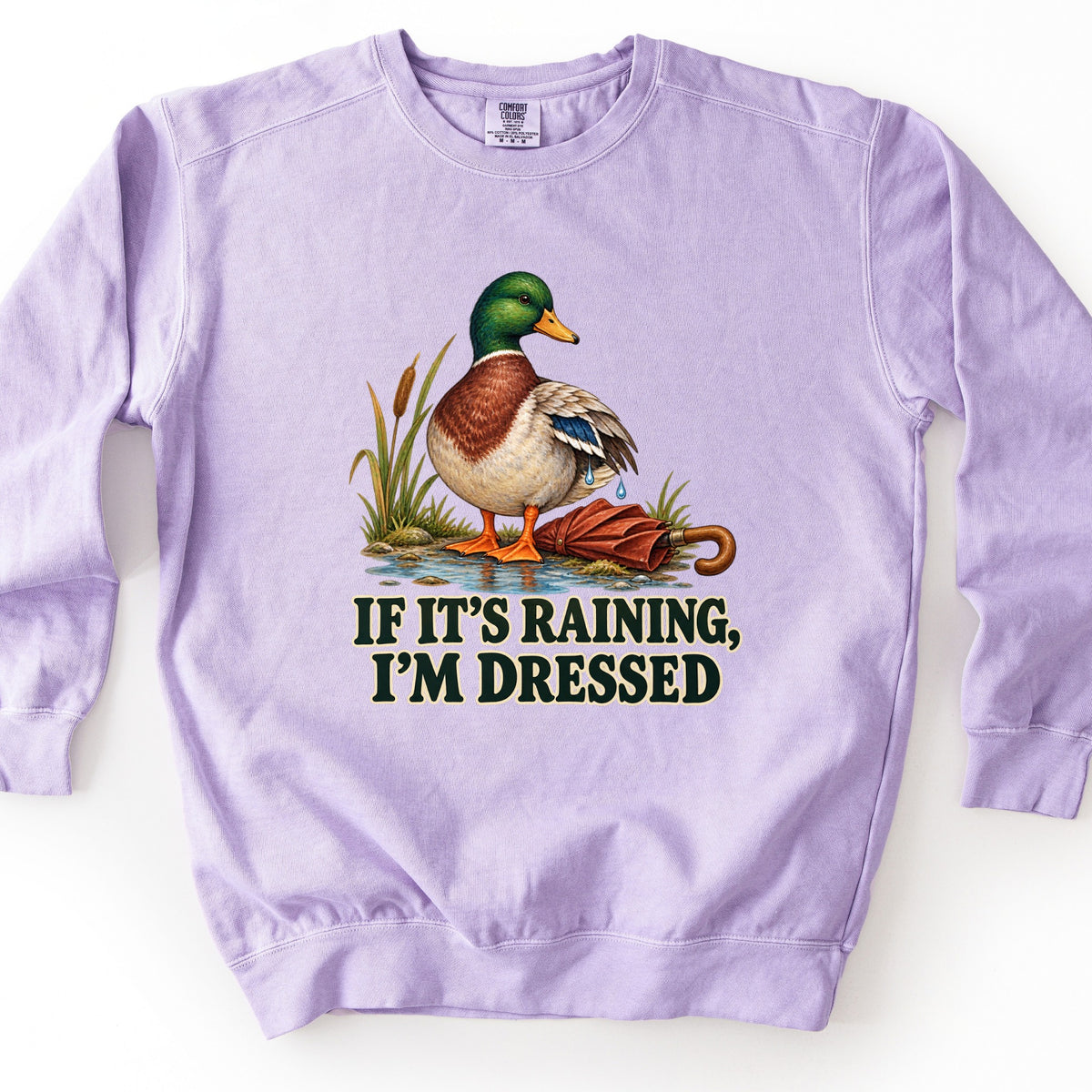 If It's Raining, I'm Dressed Comfort Colors crewneck sweatshirt in Orchid, laid flat. A mallard duck with water dripping from its wing stands in a puddle by cattails beside a closed red umbrella, above the words If It's Raining, I'm Dressed in dark green. Shown on a white background.