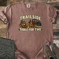 Trailside Table for Two Comfort Colors T-shirt in Espresso, laid flat. A couple in caps sits on a plaid picnic blanket with mugs and bread, pine trees and a UTV behind, between the words Trailside and Table for Two. Sleeves rolled, on wood planks with olive pants and brown boots.