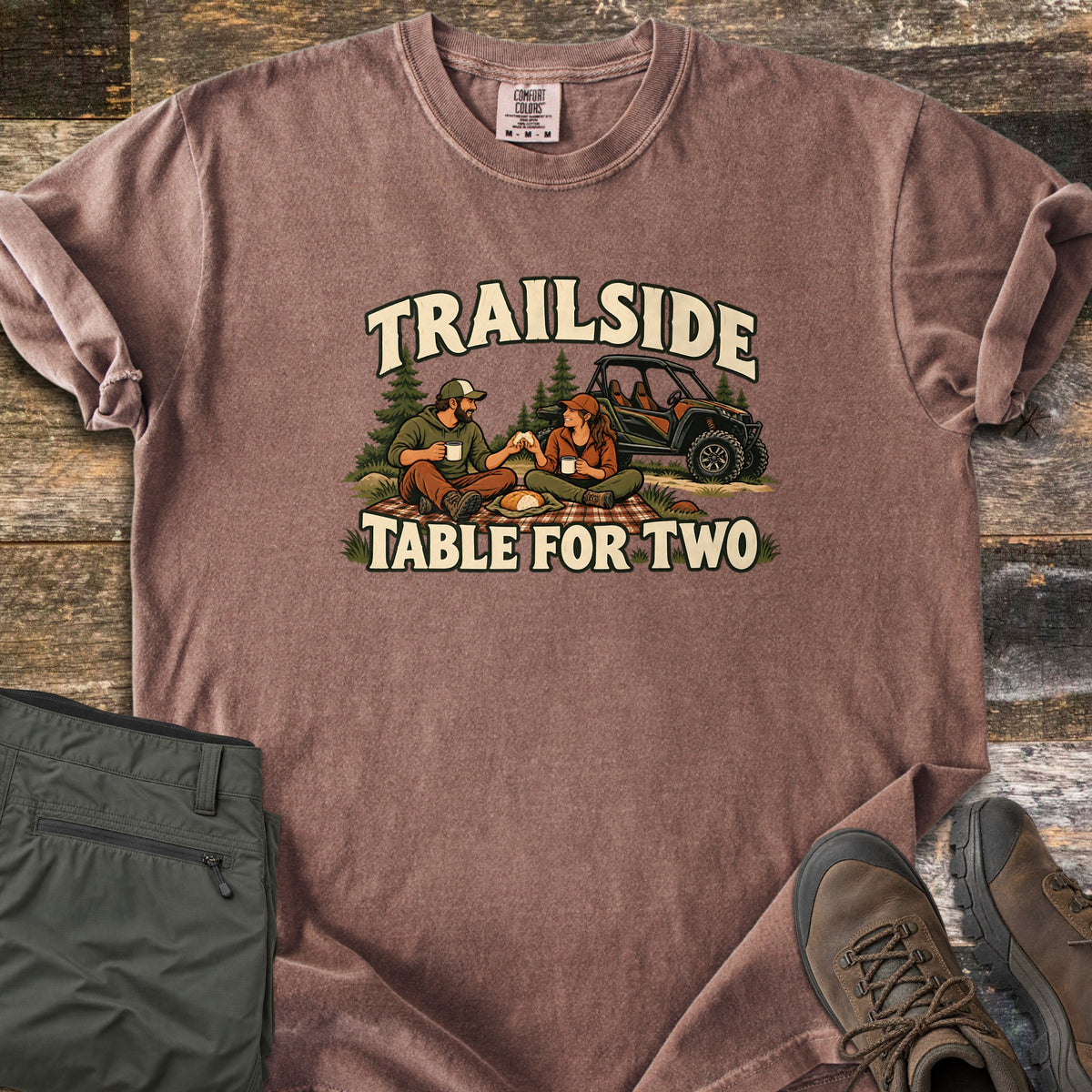 Trailside Table for Two Comfort Colors T-shirt in Espresso, laid flat. A couple in caps sits on a plaid picnic blanket with mugs and bread, pine trees and a UTV behind, between the words Trailside and Table for Two. Sleeves rolled, on wood planks with olive pants and brown boots.