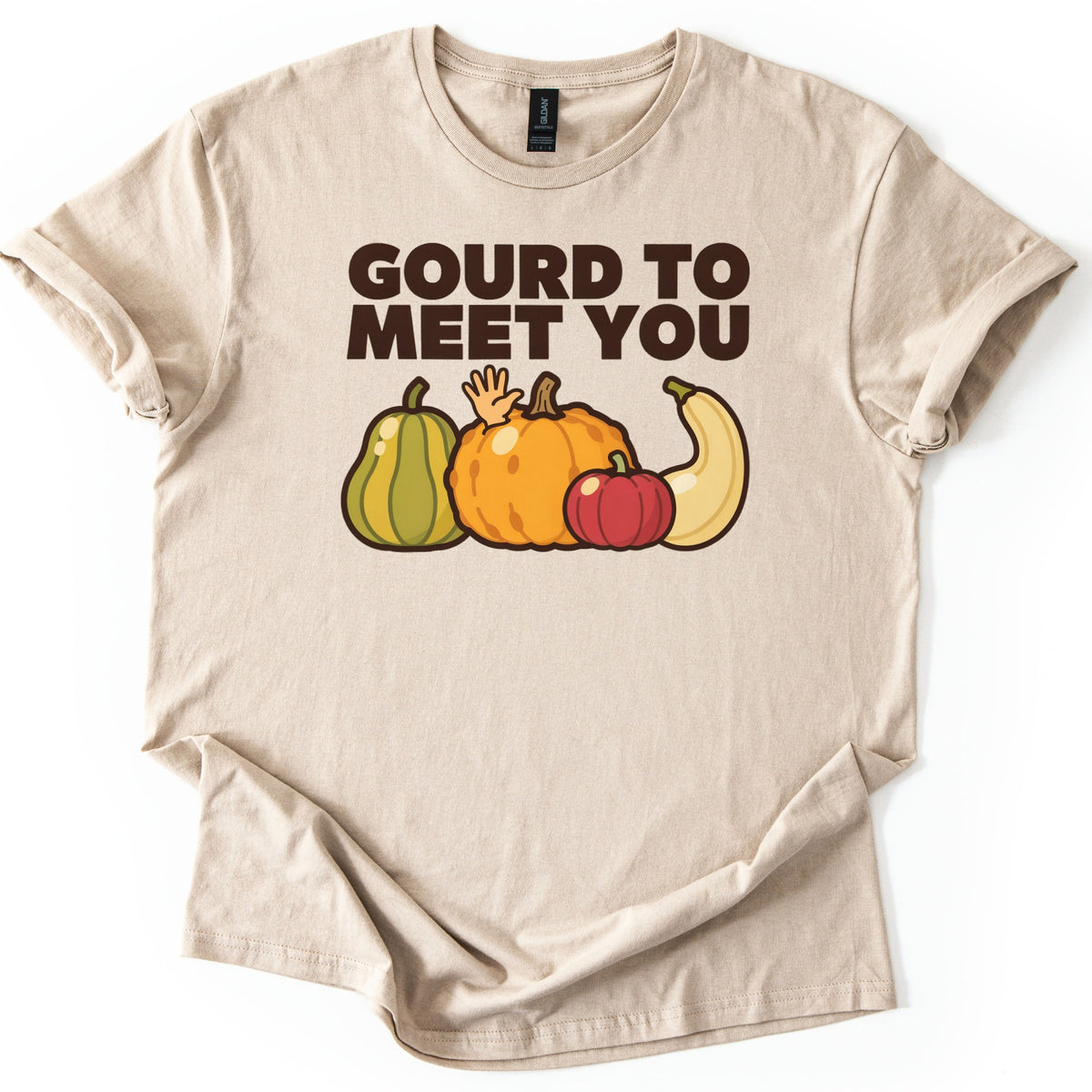 Gourd To Meet You Comfort Colors T-shirt in Sand, laid flat. Dark brown block letters read GOURD TO MEET YOU above a cartoon green squash, bumpy orange pumpkin, small red pumpkin and cream squash, with a small hand waving from behind them. Sleeves cuffed, on white.