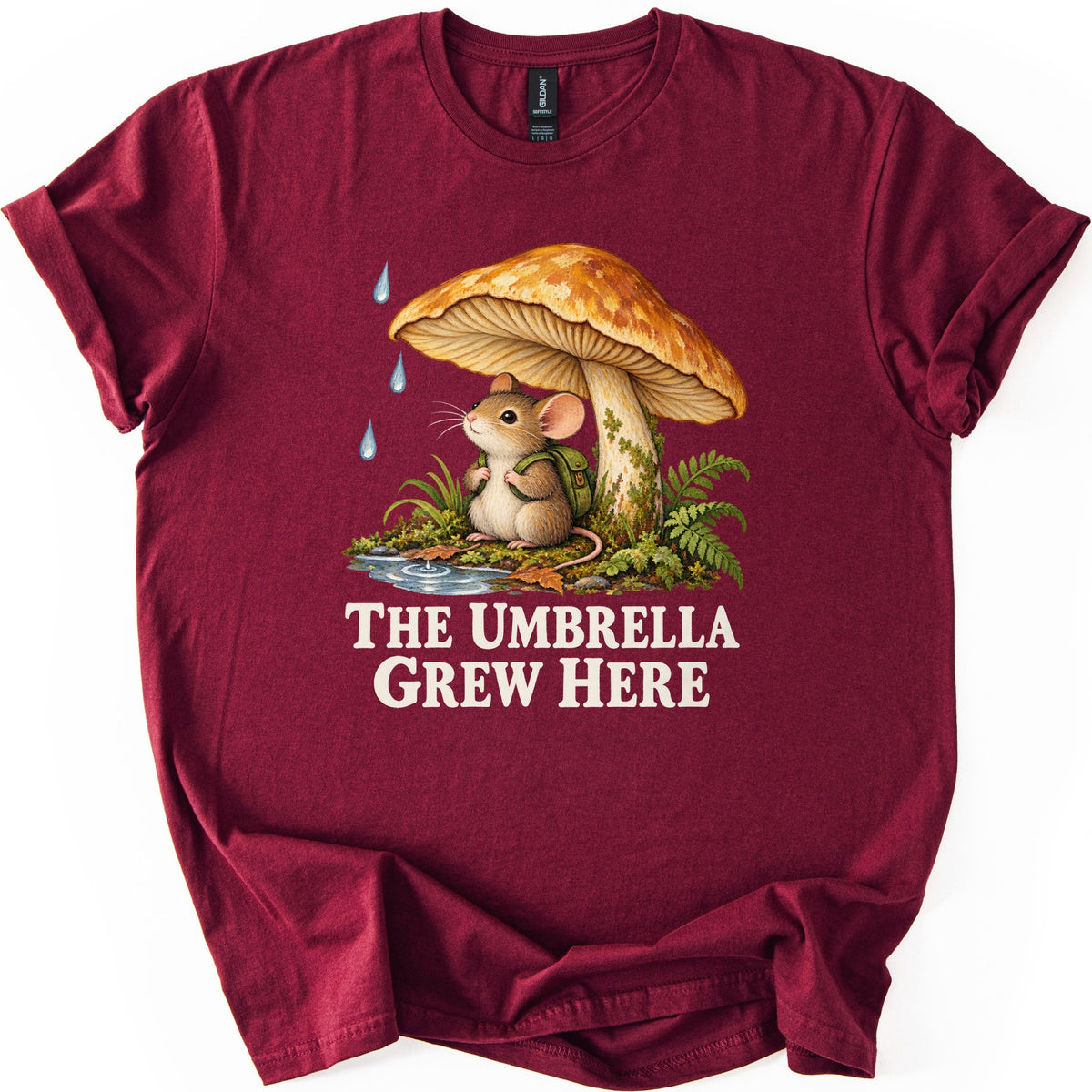 The Umbrella Grew Here Comfort Colors T-shirt in Maroon, laid flat. A mouse wearing a green backpack sits under a large orange mushroom as raindrops fall, beside ferns and a puddle, above the words The Umbrella Grew Here in cream letters. Shown on a white background.