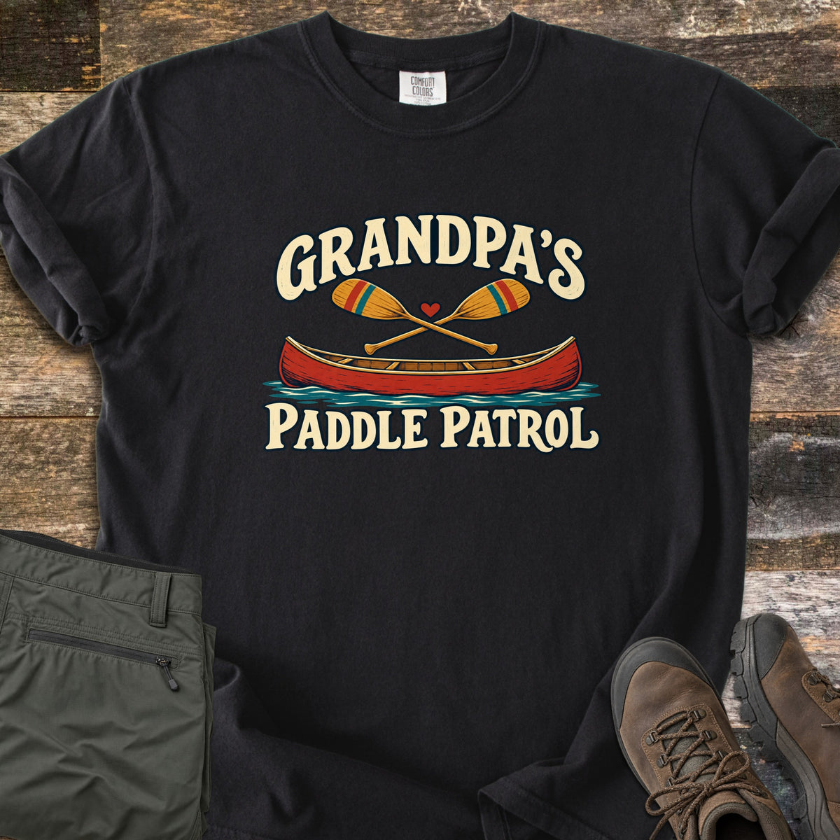 Grandpas Paddle Patrol Comfort Colors T-shirt in Black, laid flat. A red canoe on blue water with two crossed paddles and a small heart, between the words Grandpa's Paddle Patrol. Set on weathered wood beside folded olive hiking pants and brown hiking boots.