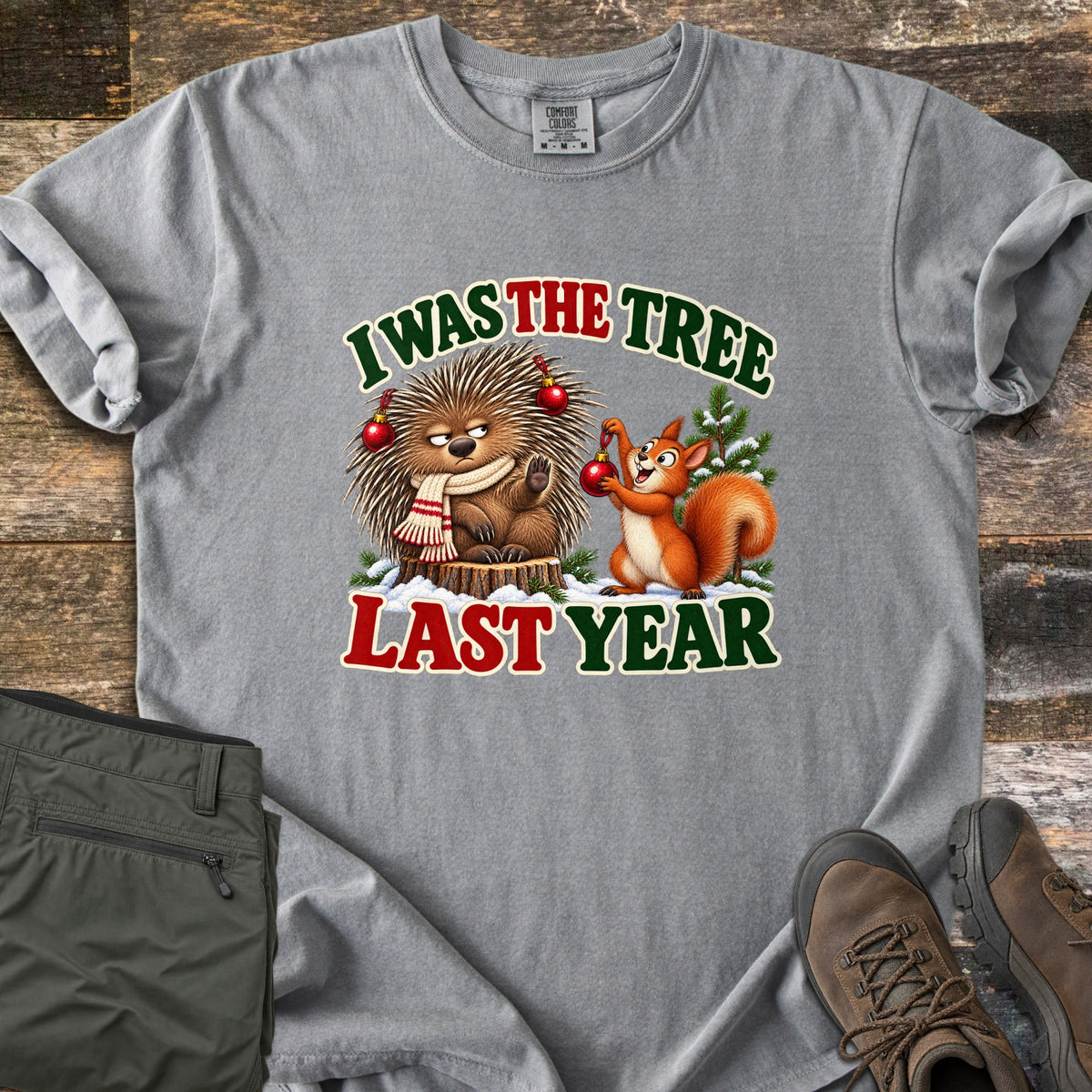 I Was The Tree Last Year Comfort Colors T-shirt in Grey, laid flat. A grumpy porcupine in a striped scarf sits on a stump with red ornaments in its quills as a smiling squirrel hangs another, between the words I Was The Tree Last Year. Olive pants and brown boots lie on wood planks.