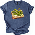 Moss Storybook Comfort Colors T-shirt in Heather Navy, laid flat. An open antique book with a red-brown cover has green moss and lichen-spotted gray rocks growing across its pages, with three slender spore stalks rising up. The shirt has rolled sleeves on a white background.