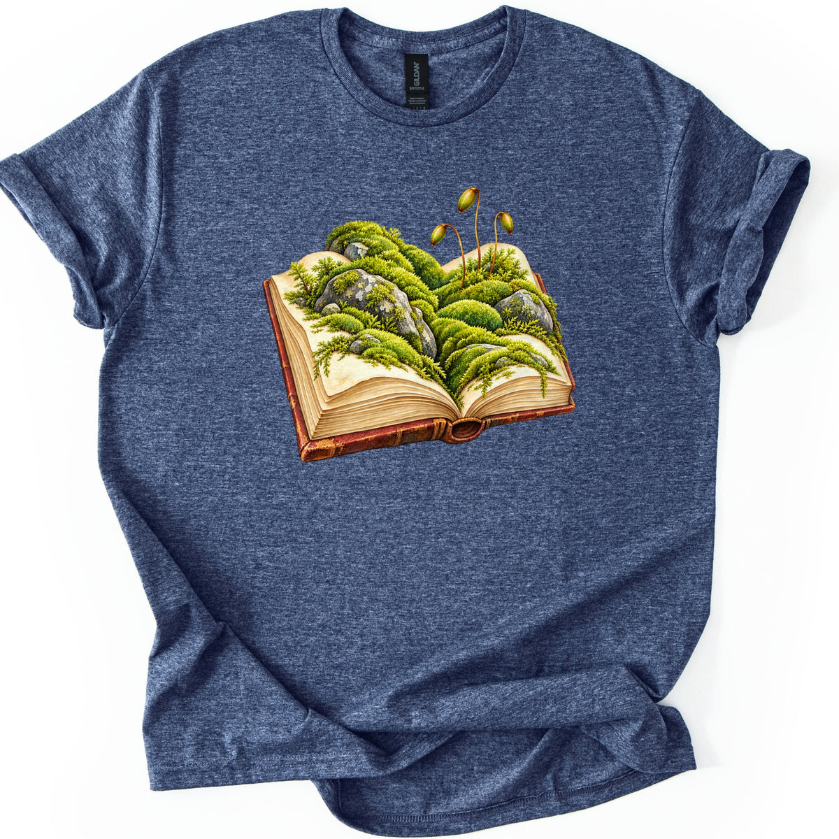 Moss Storybook Comfort Colors T-shirt in Heather Navy, laid flat. An open antique book with a red-brown cover has green moss and lichen-spotted gray rocks growing across its pages, with three slender spore stalks rising up. The shirt has rolled sleeves on a white background.