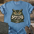 Spotted My Line Comfort Colors T-shirt in Blue Jean, laid flat. A green owl with yellow eyes sits above a winding trail between rocky ridges, with the words Spotted My Line and MTB. Olive pants and brown hiking boots lie beside it on dark wood planks.