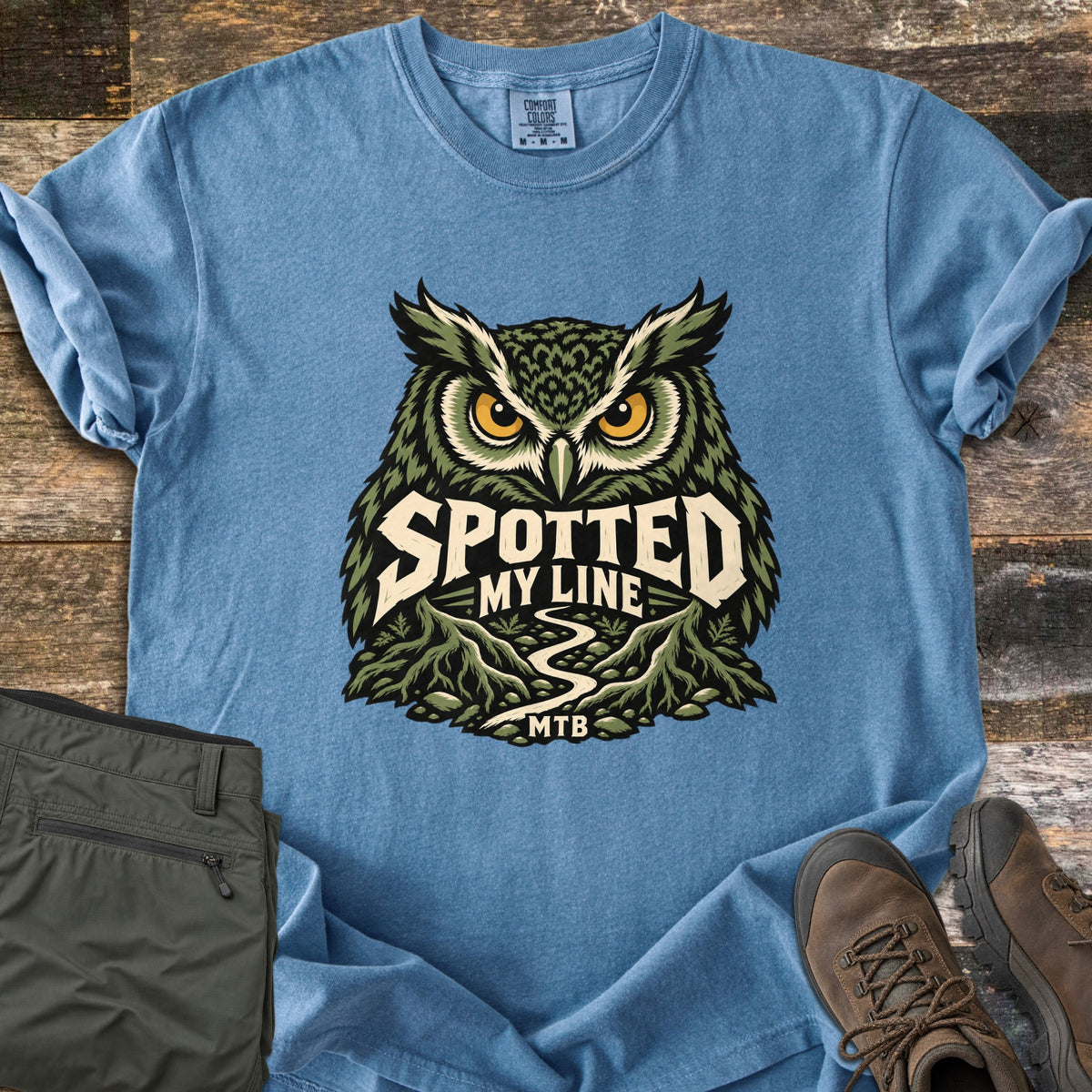 Spotted My Line Comfort Colors T-shirt in Blue Jean, laid flat. A green owl with yellow eyes sits above a winding trail between rocky ridges, with the words Spotted My Line and MTB. Olive pants and brown hiking boots lie beside it on dark wood planks.
