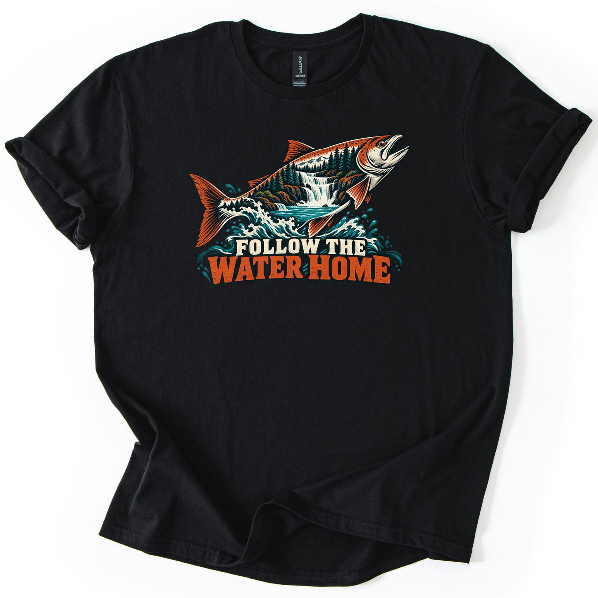 Follow the Water Home Comfort Colors T-shirt in Black, laid flat. A leaping orange salmon, its body filled with a pine forest and waterfall, rises from teal waves above the words Follow the Water Home. The shirt lies on a white background with its sleeves rolled.