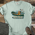 Bow Seat Bird Reporter Comfort Colors T-shirt in Bay, laid flat. A fox in a yellow life vest looks through binoculars from a green canoe as cranes fly over a lake with pines and a snowy mountain, above the words Bow Seat Bird Reporter. Olive pants and hiking boots sit on wood boards.