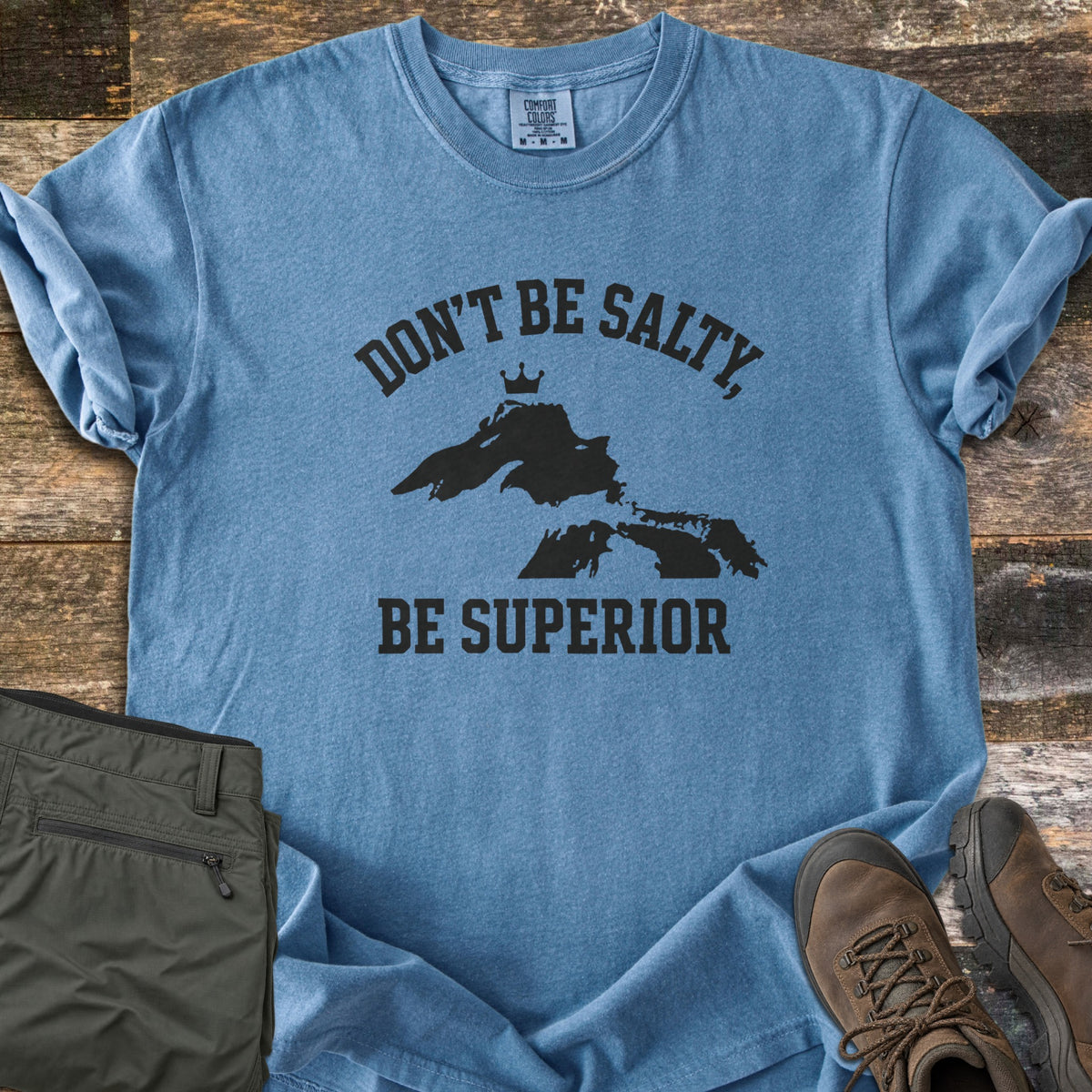 Be Superior Comfort Colors T-shirt in Blue Jean, laid flat. Black text reads Don't Be Salty, arched over a Lake Superior silhouette topped with a small crown, with Be Superior below. Sleeves rolled, on weathered wood with olive hiking pants and brown hiking boots.