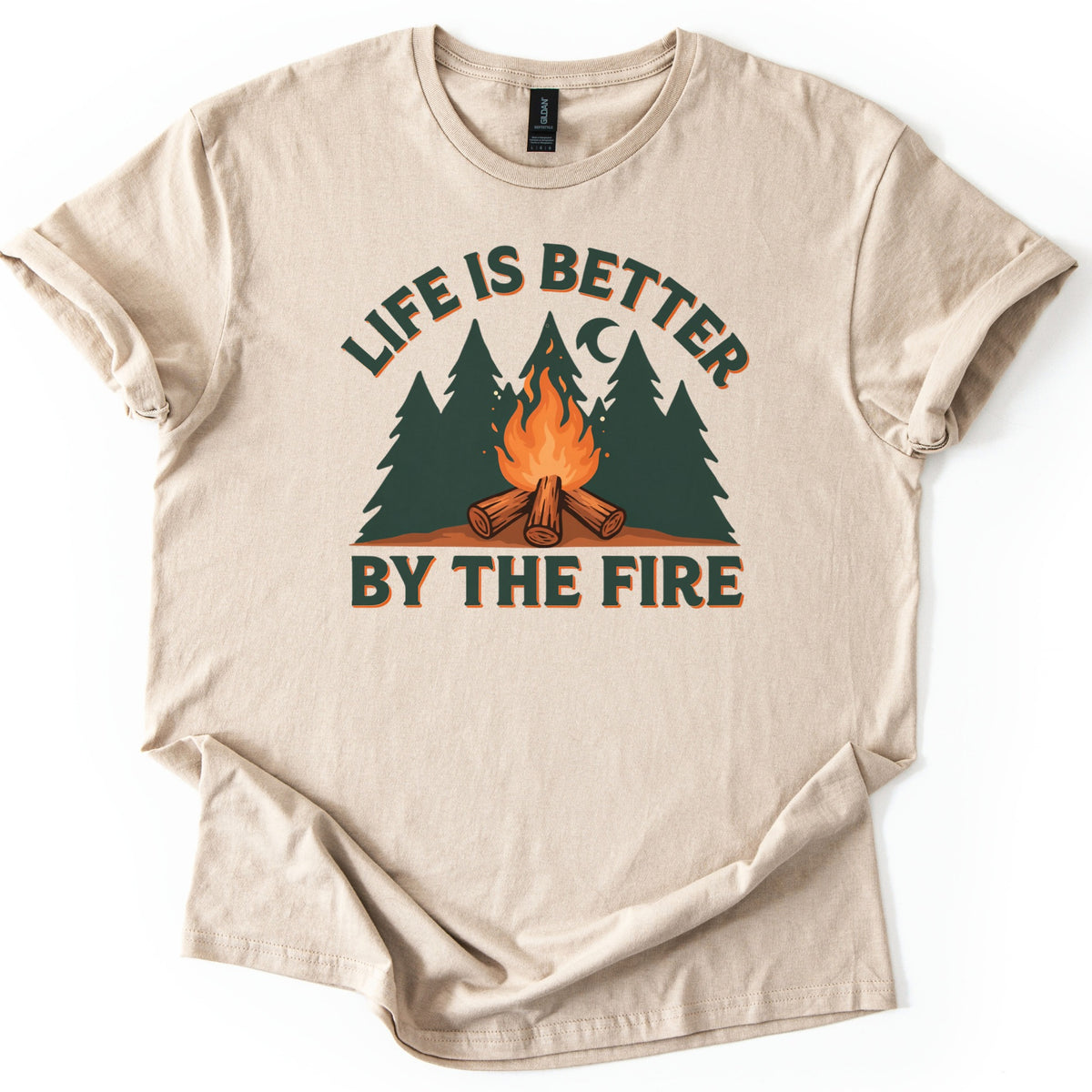 Life Is Better By The Fire Comfort Colors T-shirt in Sand, laid flat. A campfire of crossed logs burns in front of dark green pine trees under a crescent moon, with LIFE IS BETTER arched above and BY THE FIRE below in dark green letters. Cuffed sleeves, white background.