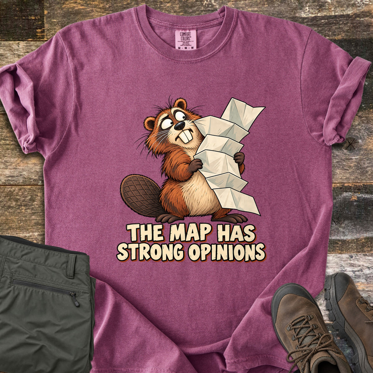 The Map Has Strong Opinions Comfort Colors T-shirt in Berry, laid flat. A cartoon beaver with wide, crossed eyes clutches a tall, unfolding paper map above the words The Map Has Strong Opinions. Sleeves are rolled, beside green hiking pants and brown hiking boots on weathered wood.