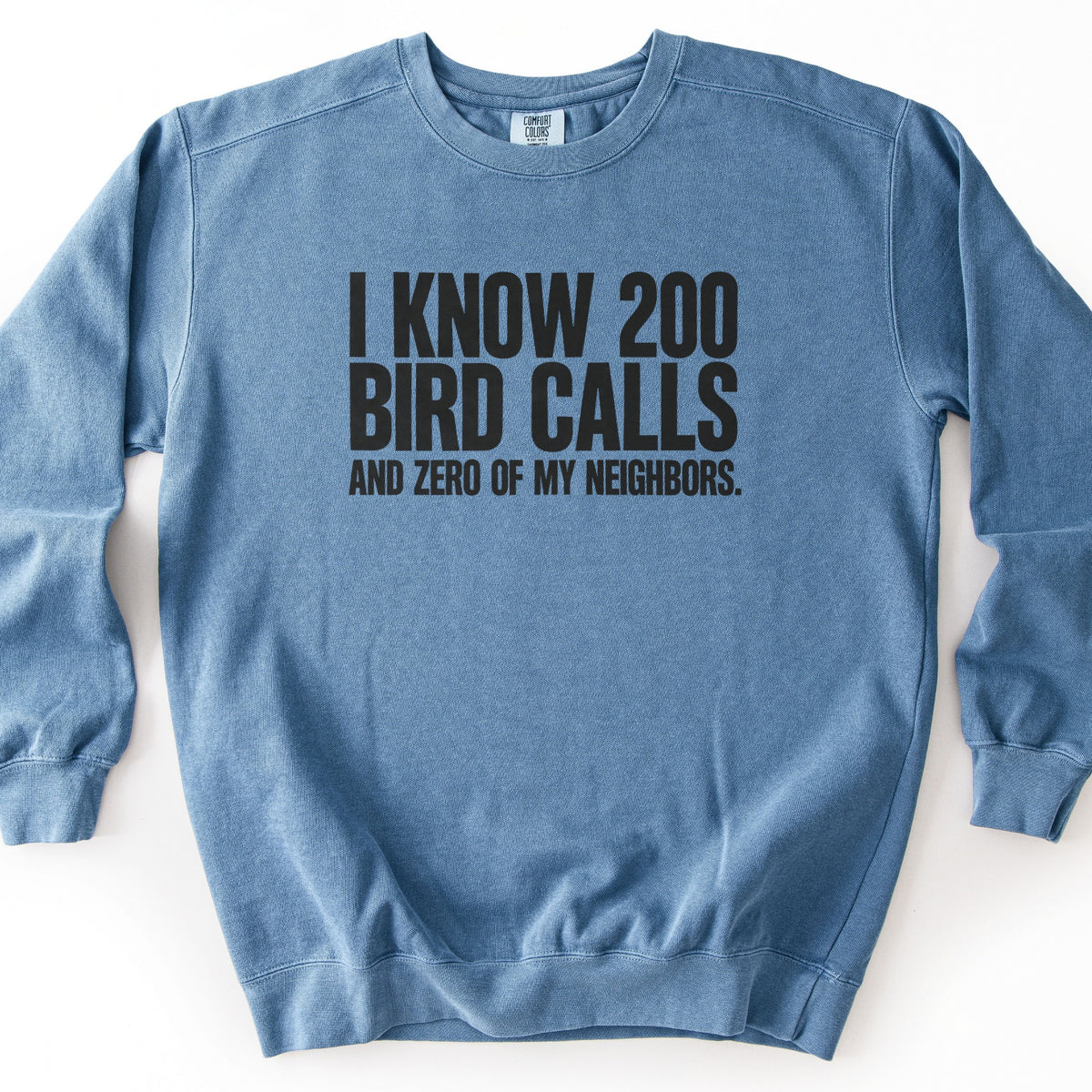I Know 200 Bird Calls and Zero of My Neighbors Comfort Colors crewneck sweatshirt in Blue Jean, laid flat. Bold black capital letters across the chest read I Know 200 Bird Calls, with And Zero of My Neighbors. in smaller letters below. The sweatshirt lies on a plain white background.