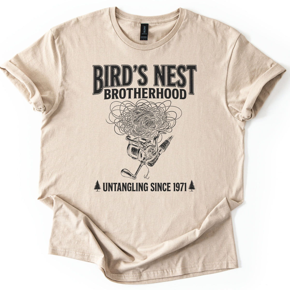 Birds Nest Brotherhood Comfort Colors T-shirt in Sand, laid flat. Black print reads Bird's Nest Brotherhood above a tangled mess of fishing line over a spinning reel, rod and hook, then Untangling Since 1971 between two pine trees. Sleeves cuffed, on a white backdrop.