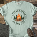 Life Is Better By The Fire Ring Comfort Colors T-shirt in Bay, laid flat. A round badge in dark green and orange shows a log campfire in front of pine trees, ringed by the words Life Is Better By The Fire. Olive hiking pants and brown hiking boots lie beside it on weathered wood planks.