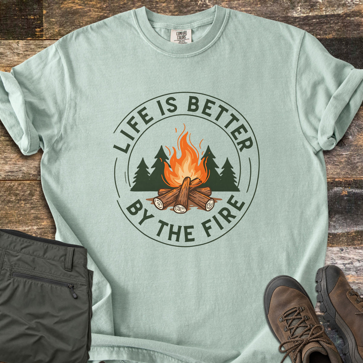 Life Is Better By The Fire Ring Comfort Colors T-shirt in Bay, laid flat. A round badge in dark green and orange shows a log campfire in front of pine trees, ringed by the words Life Is Better By The Fire. Olive hiking pants and brown hiking boots lie beside it on weathered wood planks.