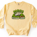 Moss Cleanup Crew Comfort Colors crewneck sweatshirt in Butter, laid flat. Green mossy letters spell Moss above a smiling white cartoon bug sweeping a brown leaf with a broom on a mound of moss, over cream letters reading Cleanup Crew. The sweatshirt lies on a white surface.