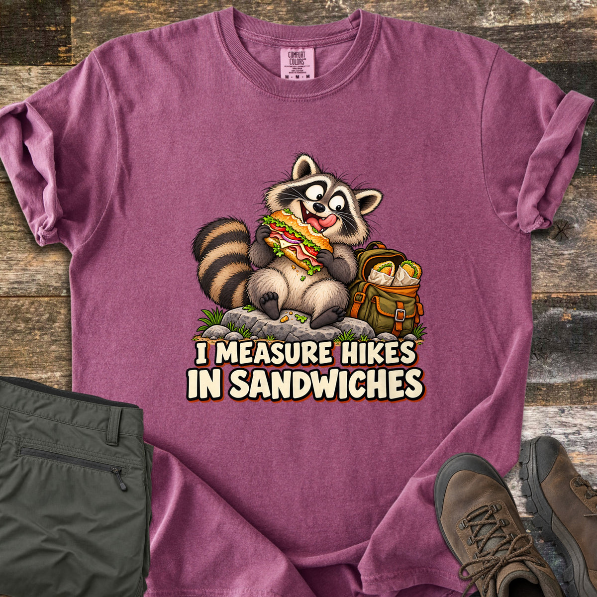 I Measure Hikes In Sandwiches Comfort Colors T-shirt in Berry, laid flat. A cartoon raccoon sits on a rock biting a big sub sandwich beside a green backpack holding wrapped burritos, above the words I Measure Hikes In Sandwiches. Hiking pants and boots lie on a wood floor beside it.