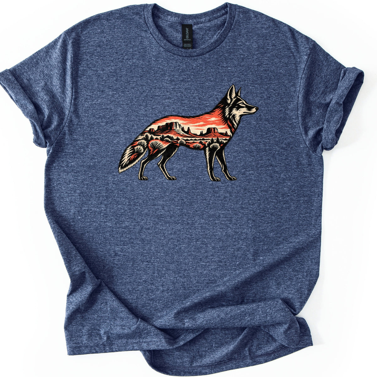 Desert Mesa Coyote Comfort Colors T-shirt in Heather Navy, laid flat. A standing coyote in profile, drawn in black and cream, has its body filled with a red desert scene of mesas, an orange sky and shrubs. No words are printed. The sleeves are rolled, on a white surface.