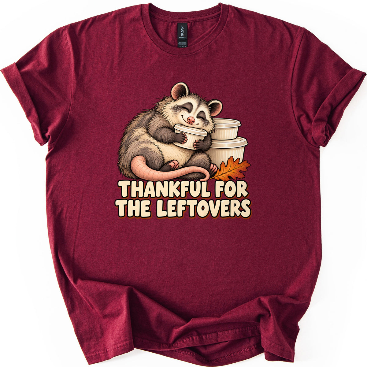 Thankful For The Leftovers Comfort Colors T-shirt in Maroon, laid flat. A plump, smiling opossum with closed eyes hugs a white food container beside a stack of more containers and an orange oak leaf, above the words Thankful For The Leftovers. Sleeves are cuffed on a white surface.