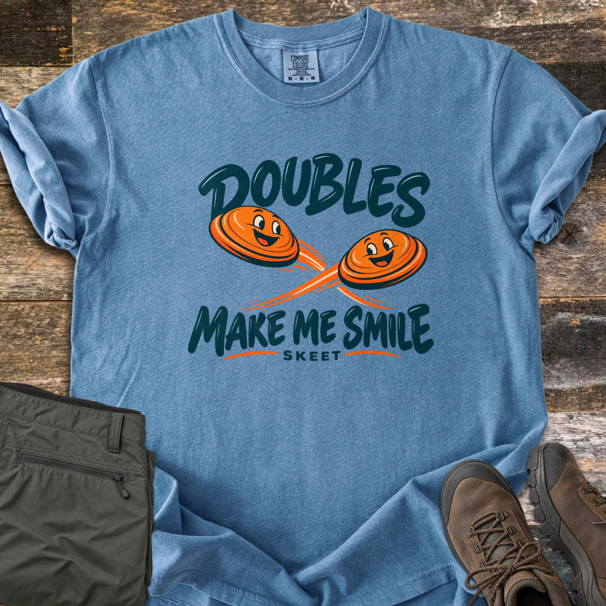 Doubles Make Me Smile Comfort Colors T-shirt in Blue Jean, laid flat. Two smiling orange clay targets fly on crossing paths between the words Doubles Make Me Smile, with Skeet printed below. Folded olive pants and brown hiking boots sit beside it on weathered wood planks.