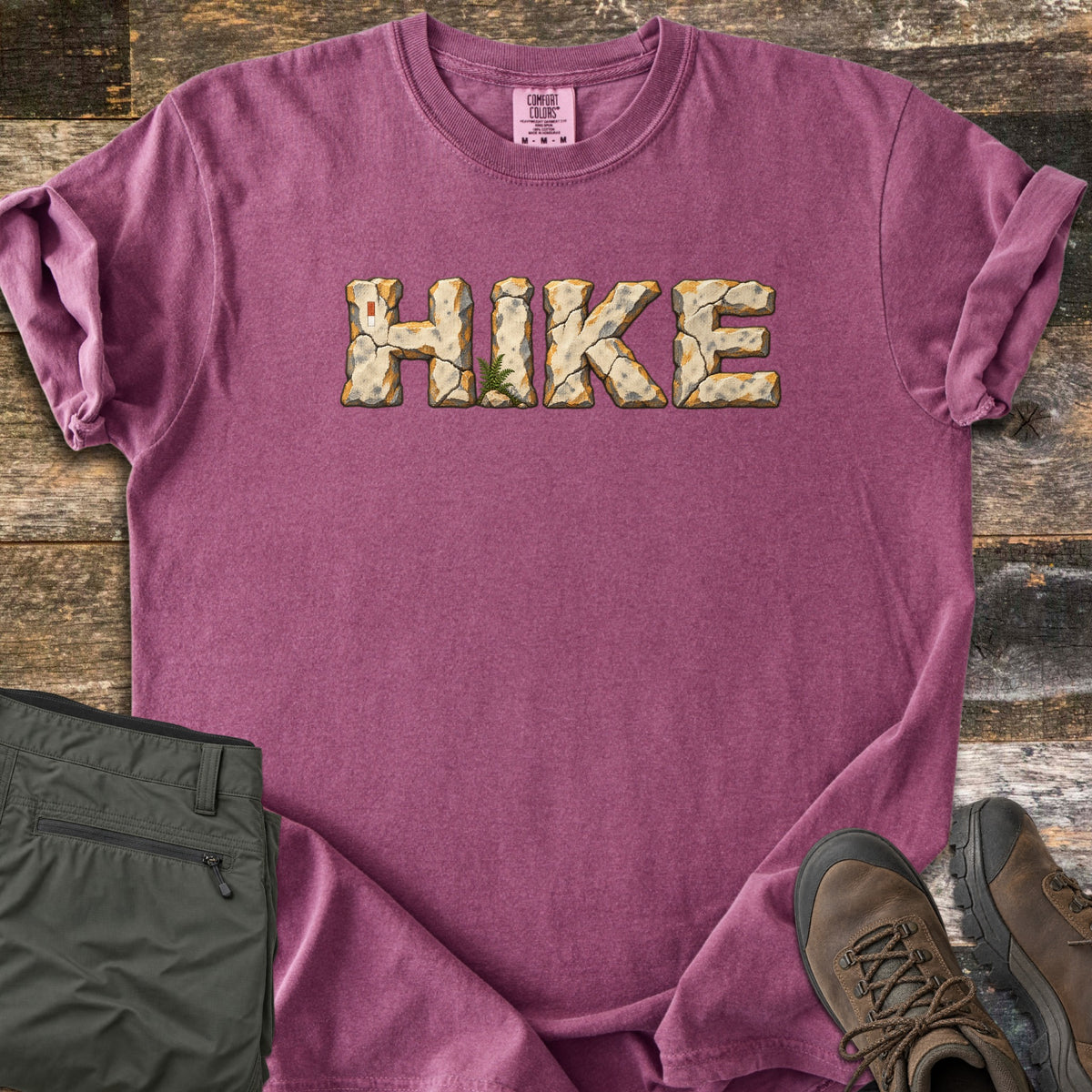 Hike Comfort Colors T-shirt in Berry, laid flat. The word HIKE is printed across the chest in cracked stone letters with orange edges, a small fern at the base of the I. Olive hiking pants and brown hiking boots lie beside it on weathered wood planks.