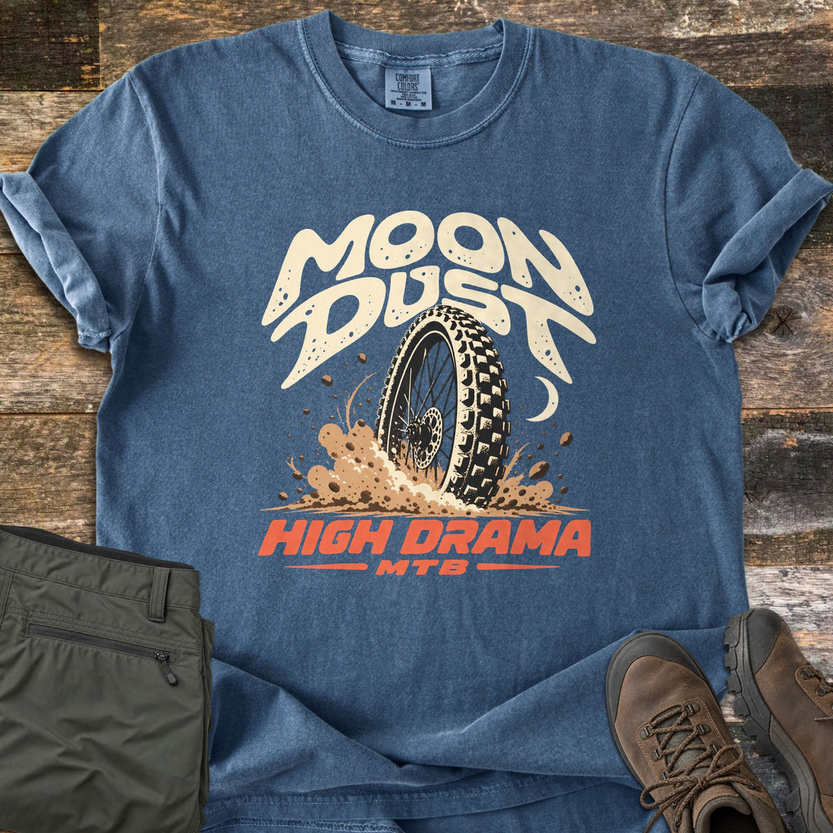 Moon Dust High Drama Comfort Colors T-shirt in Denim, laid flat. Cream cratered letters reading Moon Dust arch over a knobby bike tire spraying dirt beside a crescent moon, with High Drama MTB in orange below. Sleeves rolled, on wood planks with olive pants and brown hiking boots.